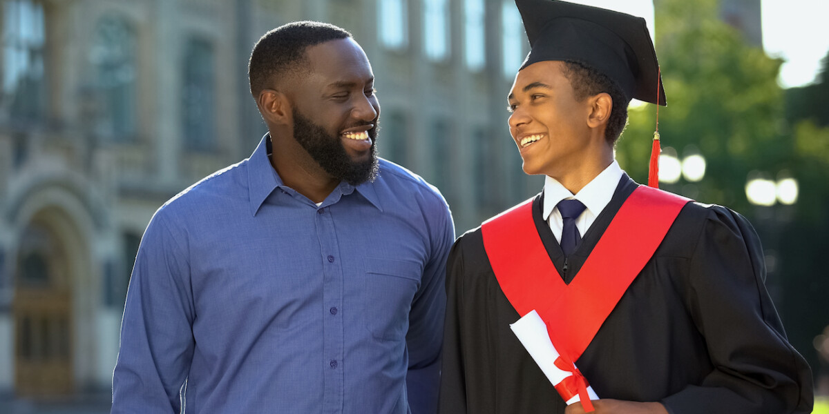 Smiling black father embracing graduating son with diploma education degree